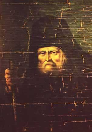 Portrait of the elderly Seraphim of Sarov in a monastic klobuk and mantle, holding a staff, his lit face set against a very dark ground, the paint covered in fine cracks.